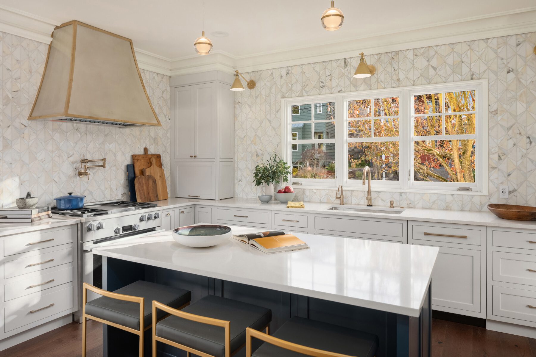 Modern kitchen with white cabinets, marble backsplash, island seating, gold accents, and large windows showing autumn trees outside. Open book and bowl on the island.