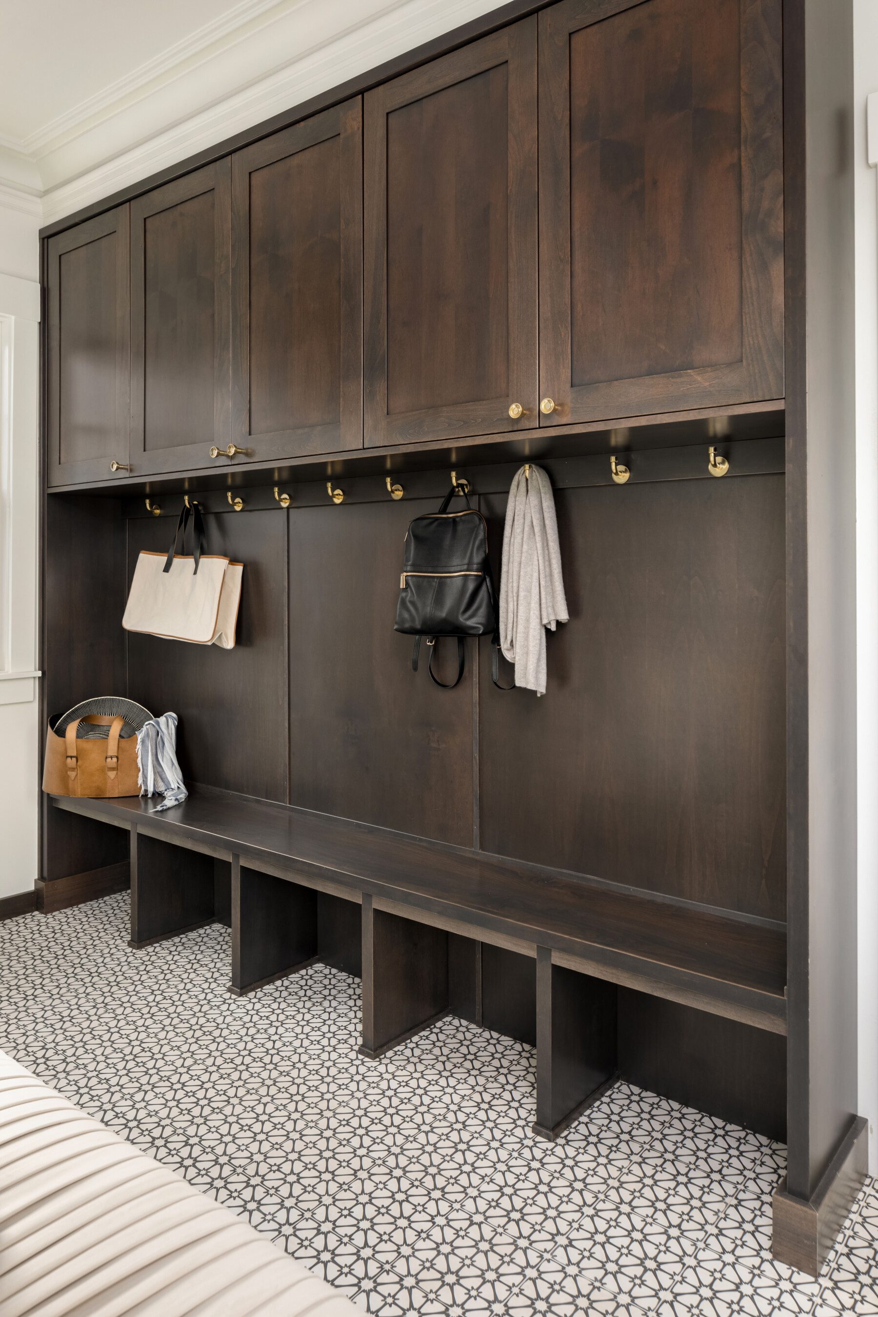 A mudroom features dark wood cabinets, brass hooks holding bags and a scarf, open bench seating with cubbies below, and patterned tile flooring.