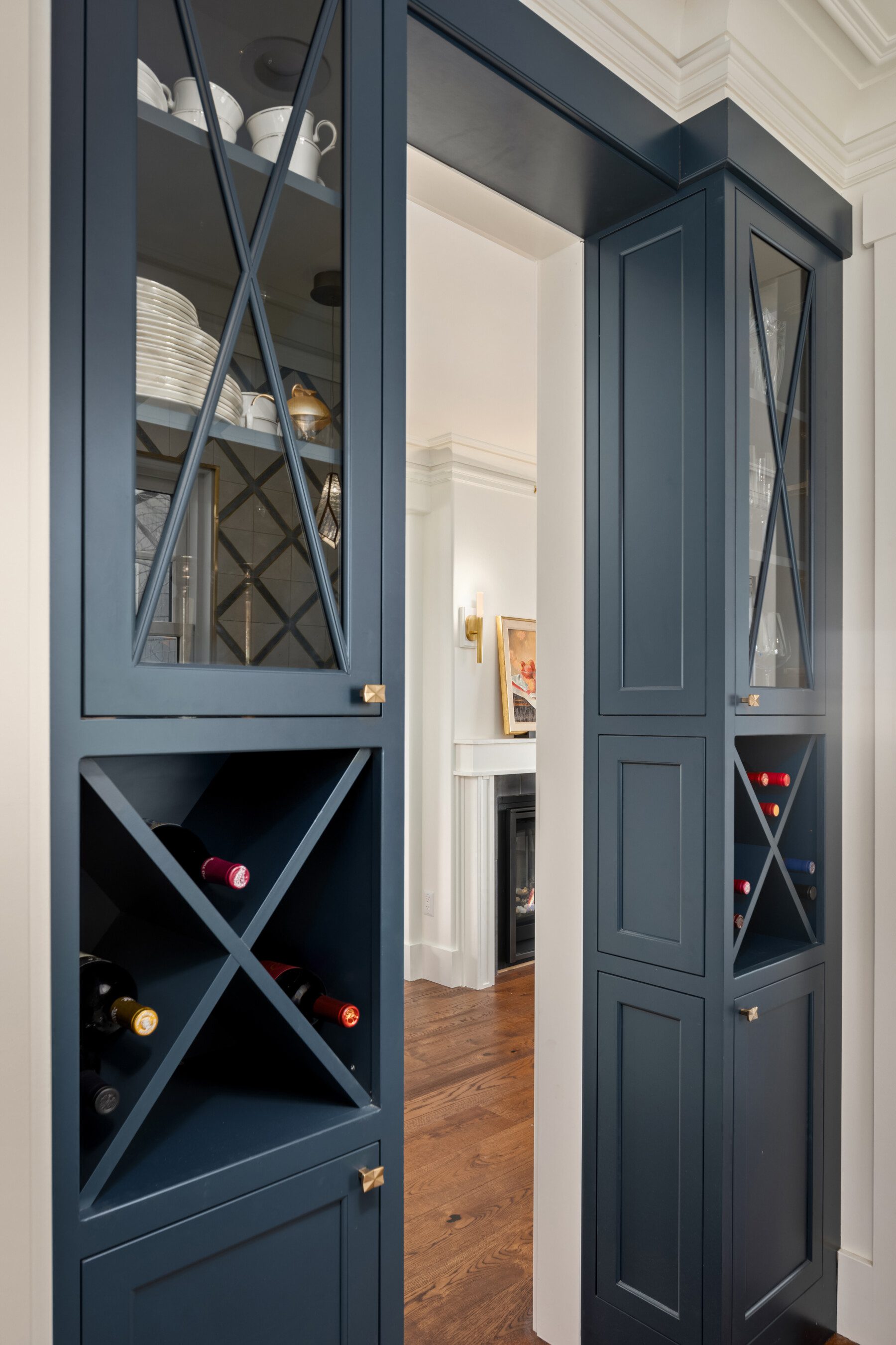 Built-in navy blue cabinetry with glass-front doors, shelves holding dishes, X-shaped wine storage, and a view into a living area with a fireplace and wall sconce.