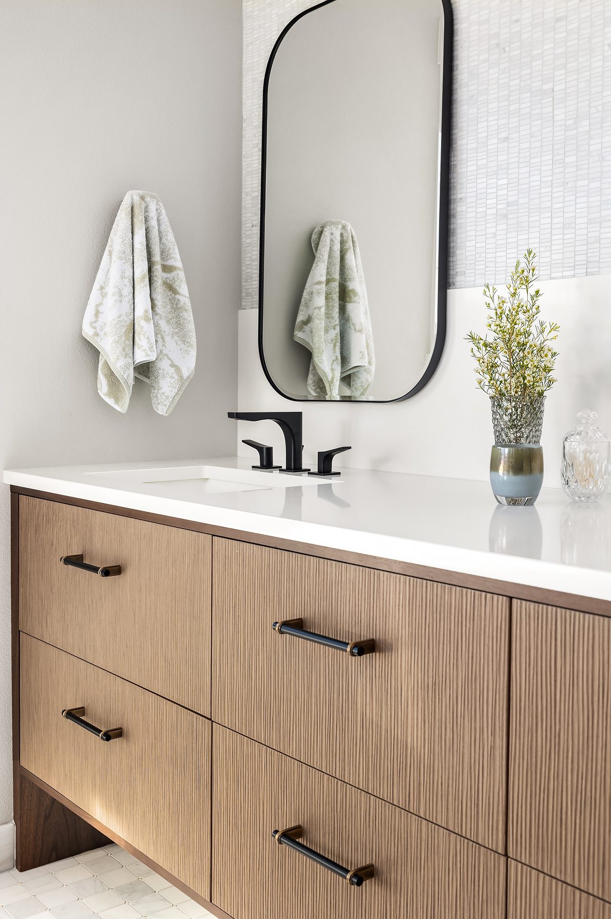 Modern bathroom vanity with wooden drawers, black fixtures, a rectangular mirror, a towel on the wall, and a vase with greenery on a white countertop.