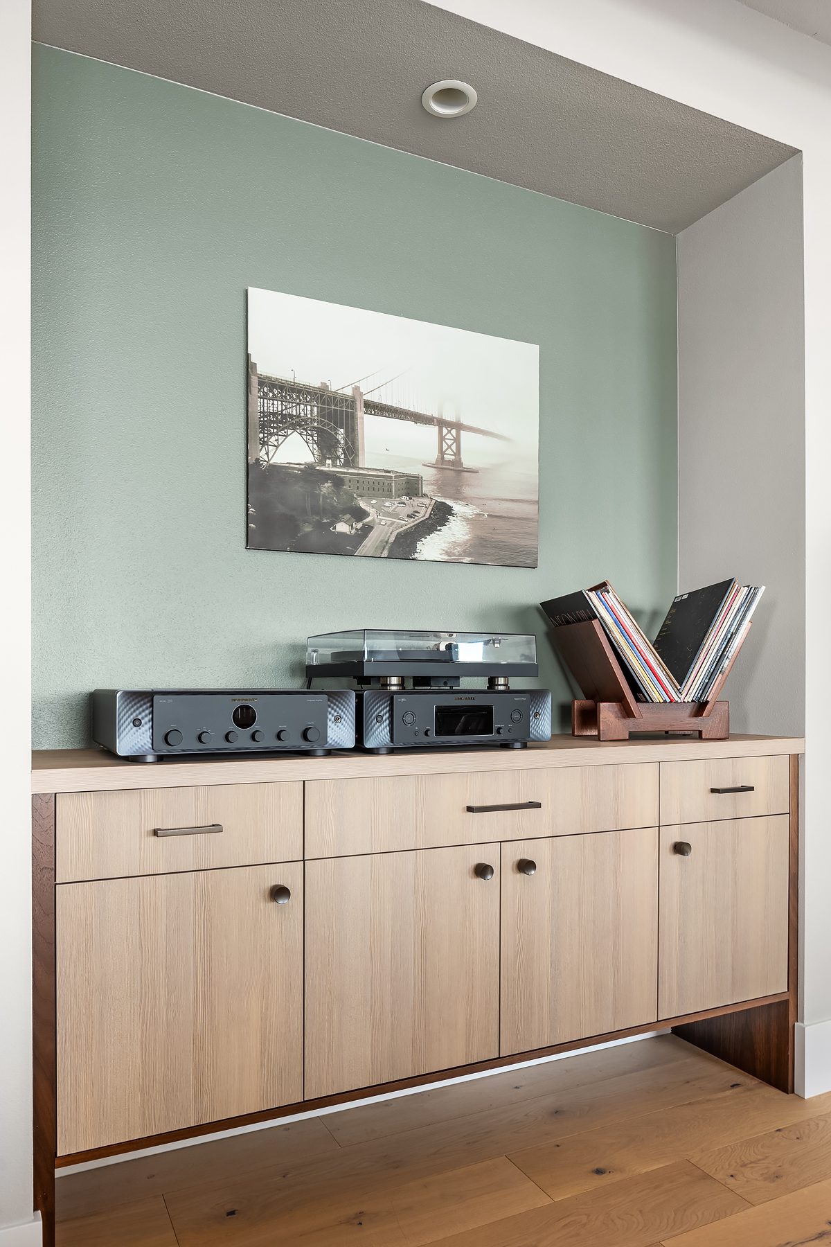 A wooden cabinet with audio equipment, a turntable, and vinyl records sits beneath a framed photo of a bridge on a green accent wall.