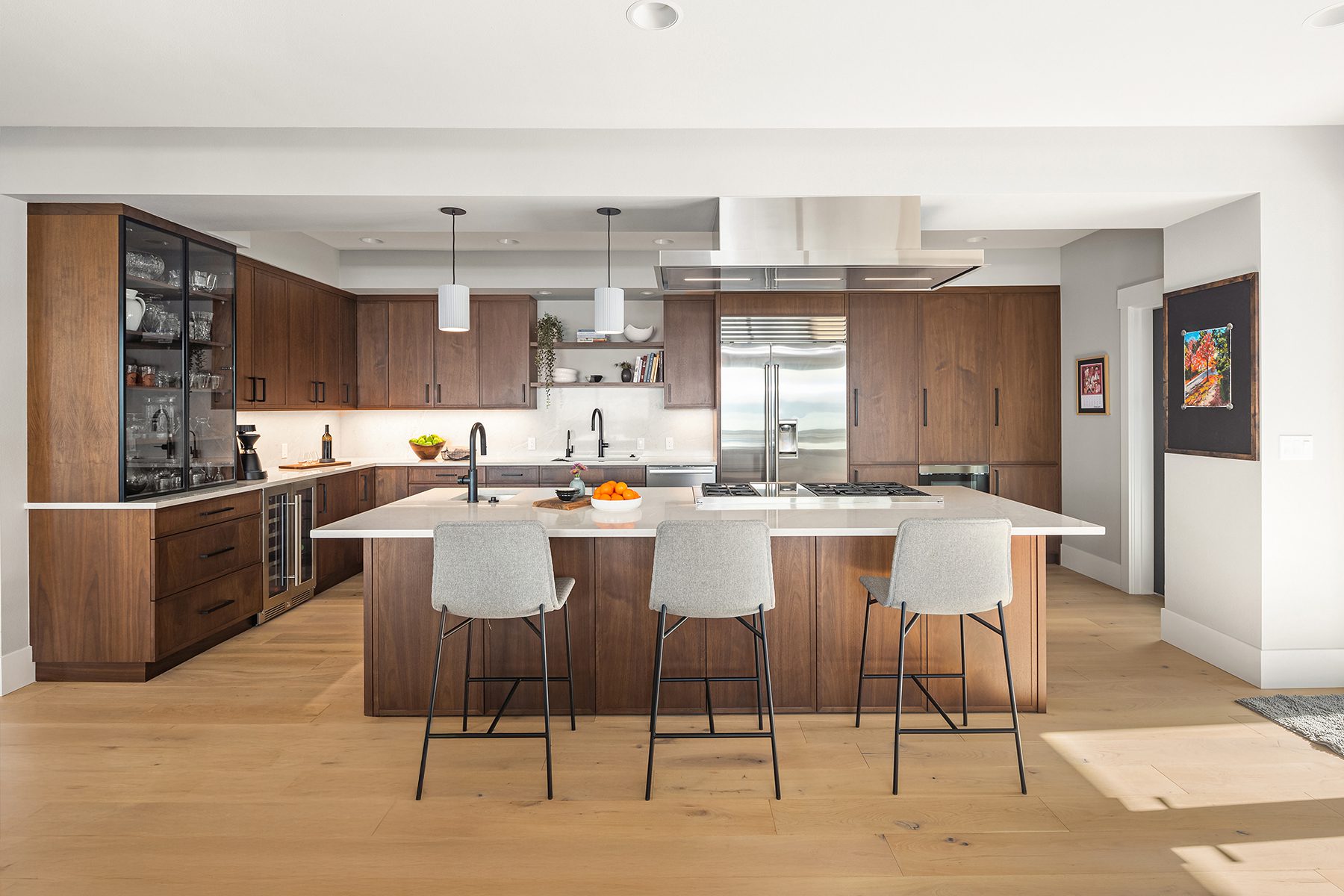 Modern kitchen with wooden cabinets, stainless steel appliances, a large island with three gray barstools, pendant lights, and light wood flooring.