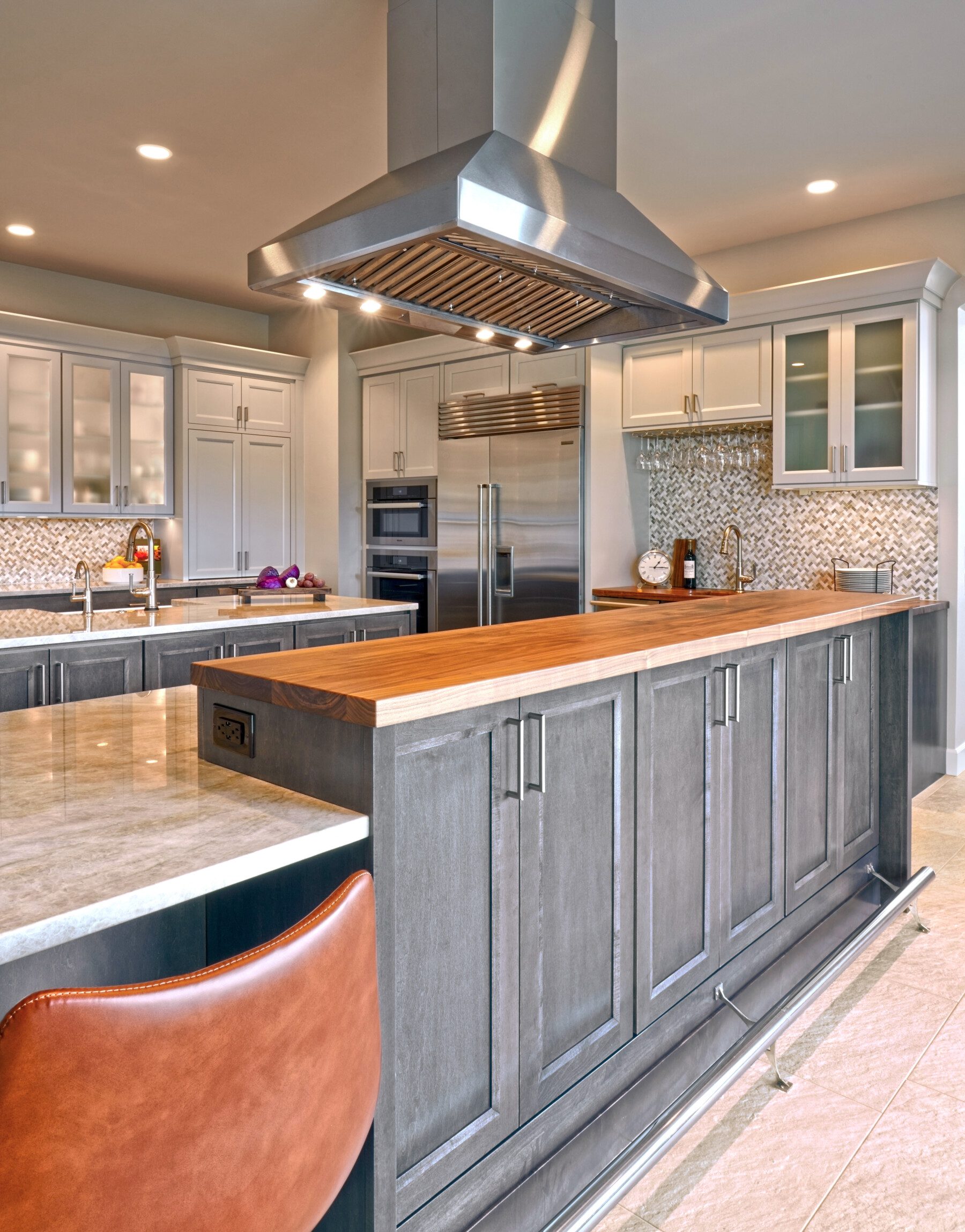 Modern kitchen with stainless steel appliances, a large island with wood countertop, gray cabinets, leather barstool, and mosaic tile backsplash.