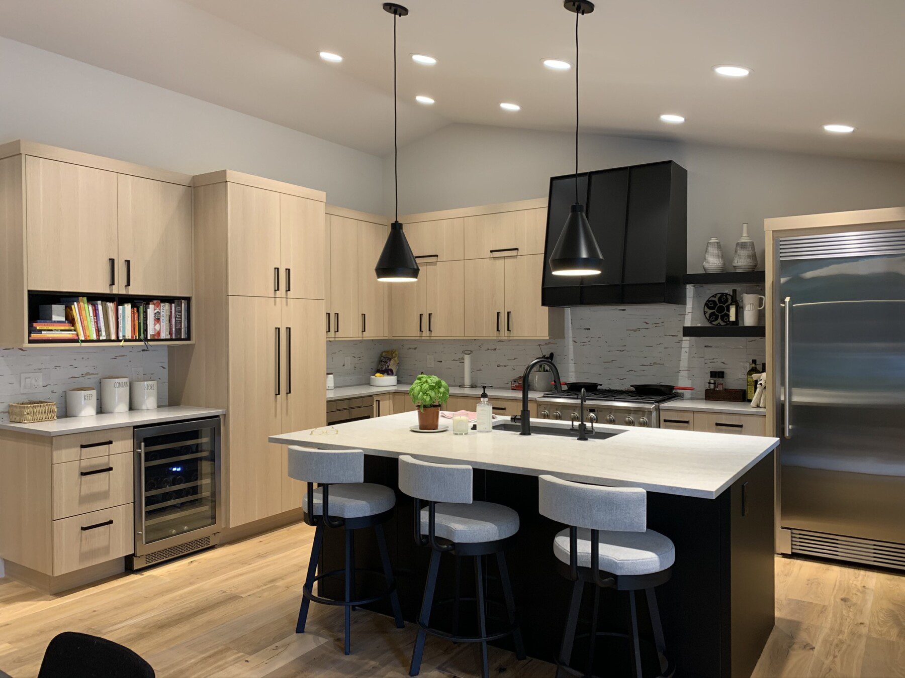 Modern kitchen with light wood cabinets, black accents, a center island with three stools, pendant lights, built-in appliances, and a stainless steel refrigerator.