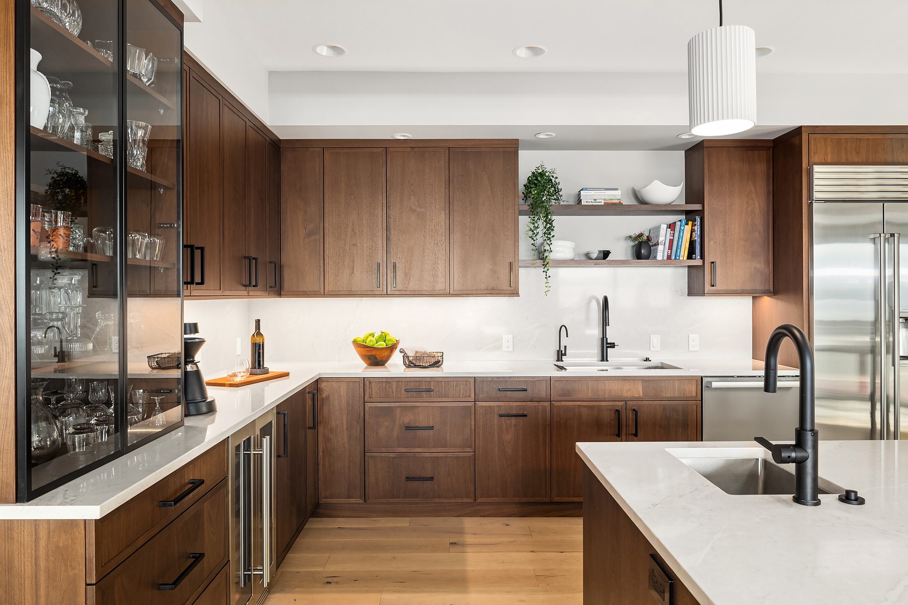 Modern kitchen with brown wooden cabinets, white countertops, stainless steel appliances, a kitchen island with sink, and open shelving with dishes and plants.