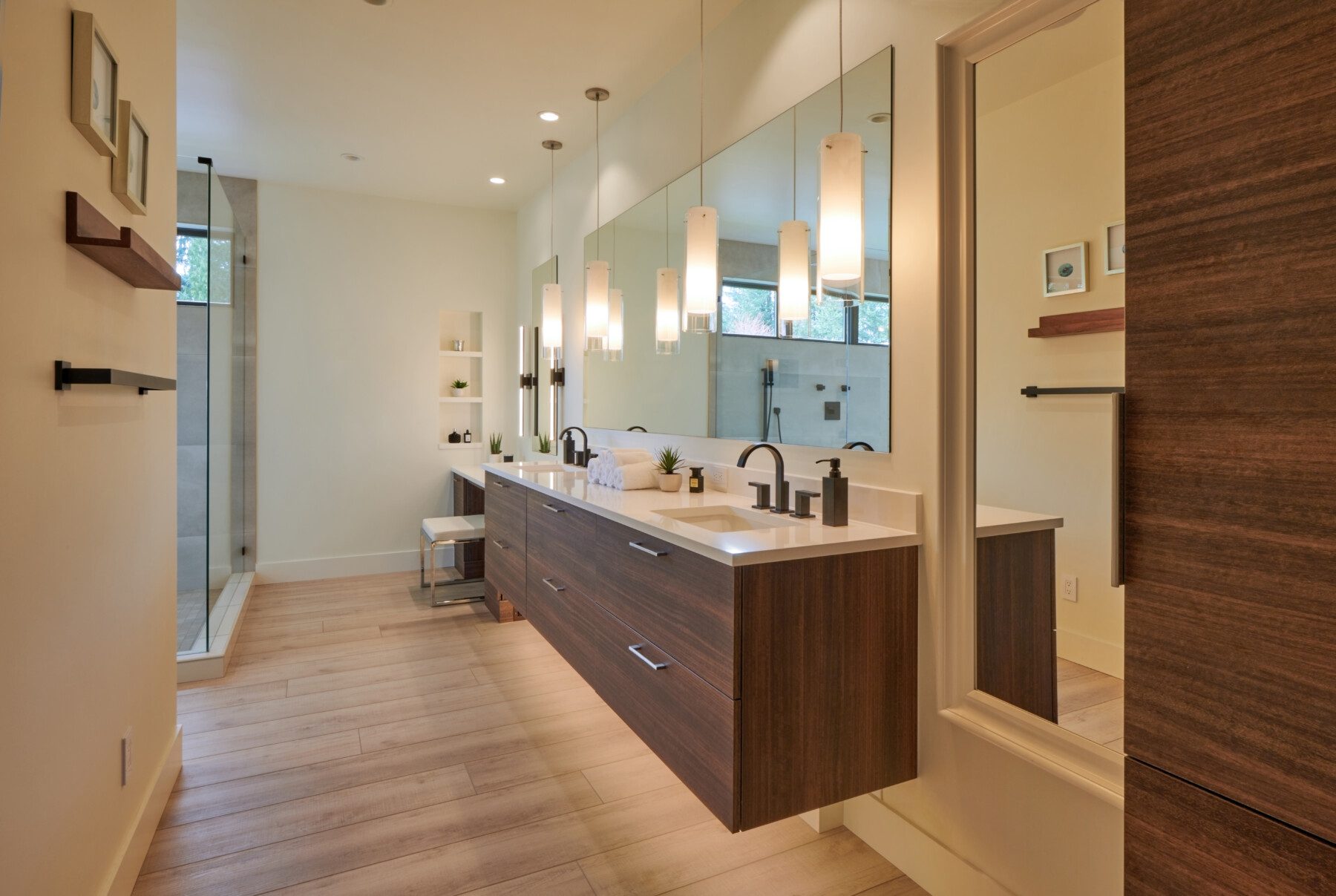 Modern bathroom with double sinks, wood cabinetry, large mirrors, pendant lights, a walk-in shower, and light wood flooring.