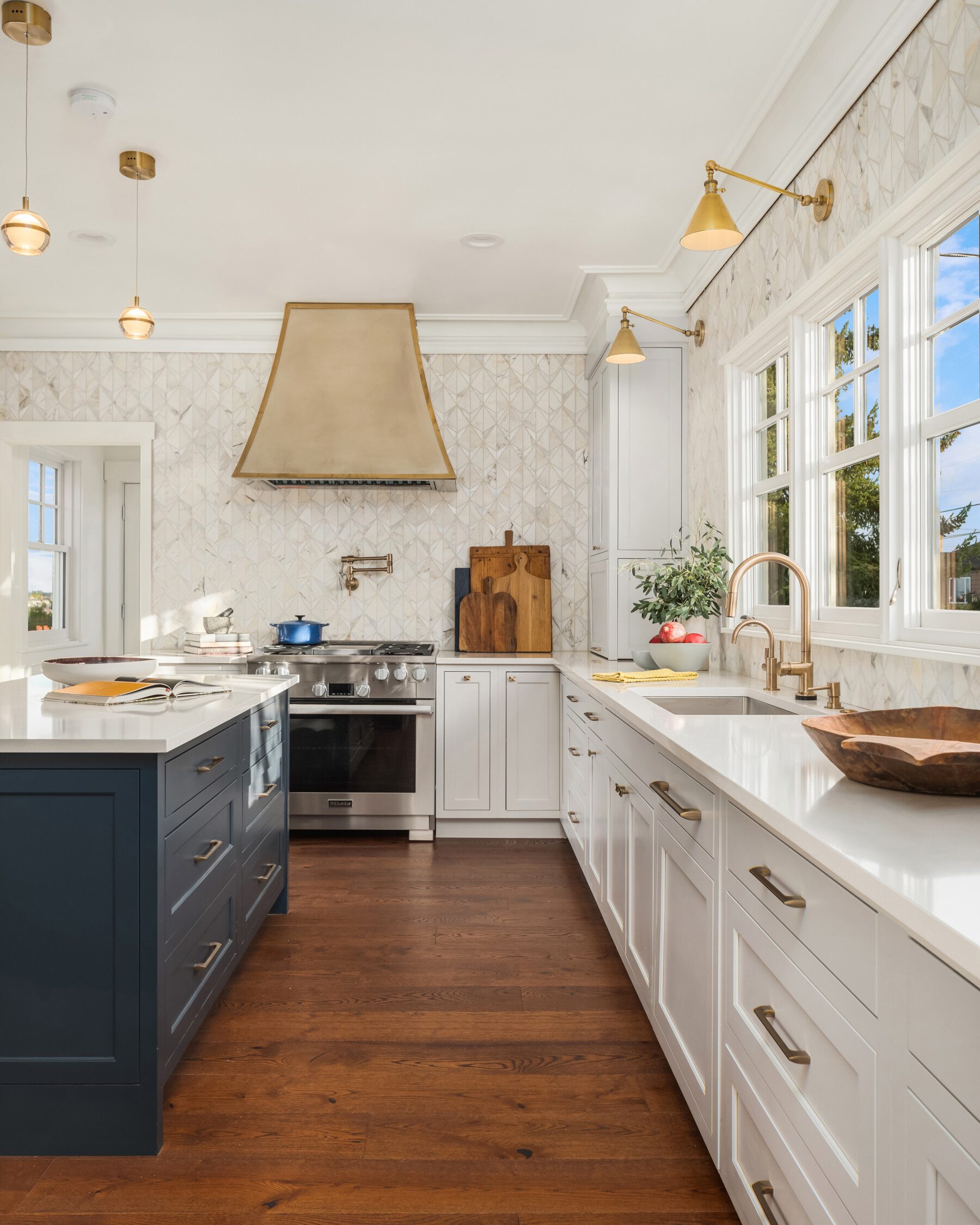 Modern kitchen with white cabinets, wood flooring, gold fixtures, a navy island, stainless steel stove, brass range hood, and large windows letting in natural light.