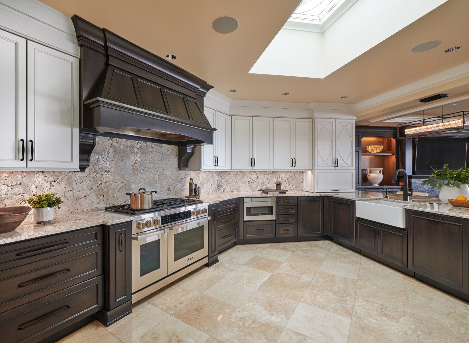 Modern kitchen with dark lower cabinets, white upper cabinets, stainless steel appliances, a farmhouse sink, and beige tiled floor under a skylight.