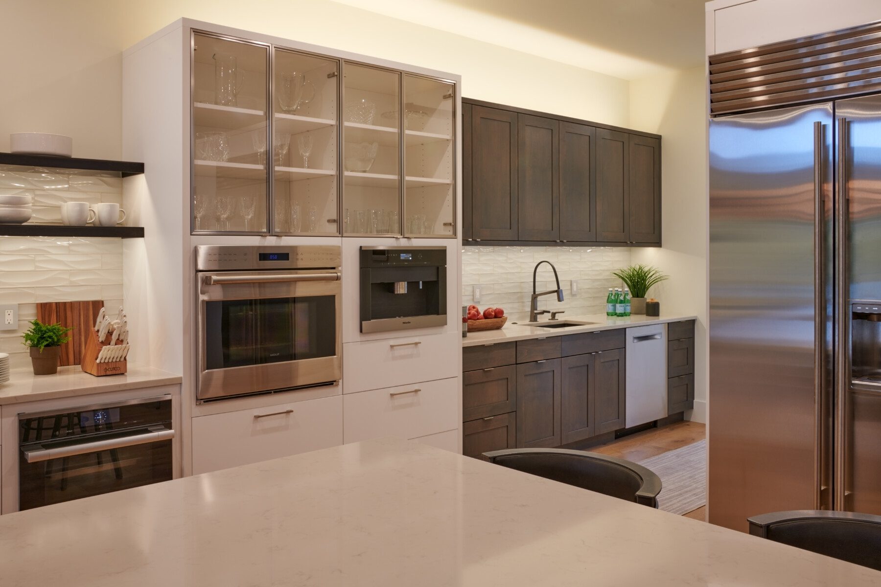 Modern kitchen with stainless steel appliances, glass-front cabinets, dark wood lower cabinets, a white countertop, and a backsplash with textured white tiles.
