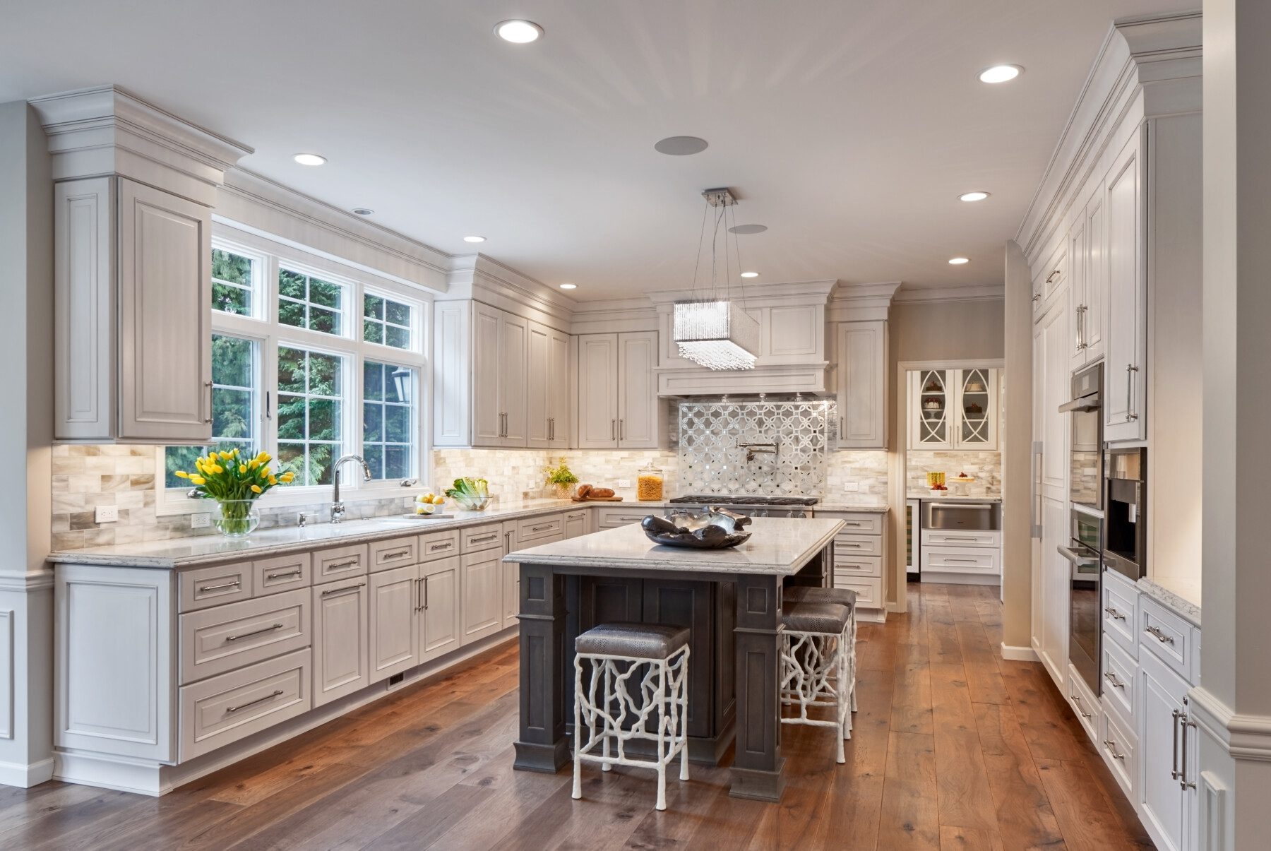 Modern kitchen with white cabinetry, marble countertops, a central island with stools, stainless steel appliances, wood floors, and decorative lighting. Flowers and fruit bowl accent the space.