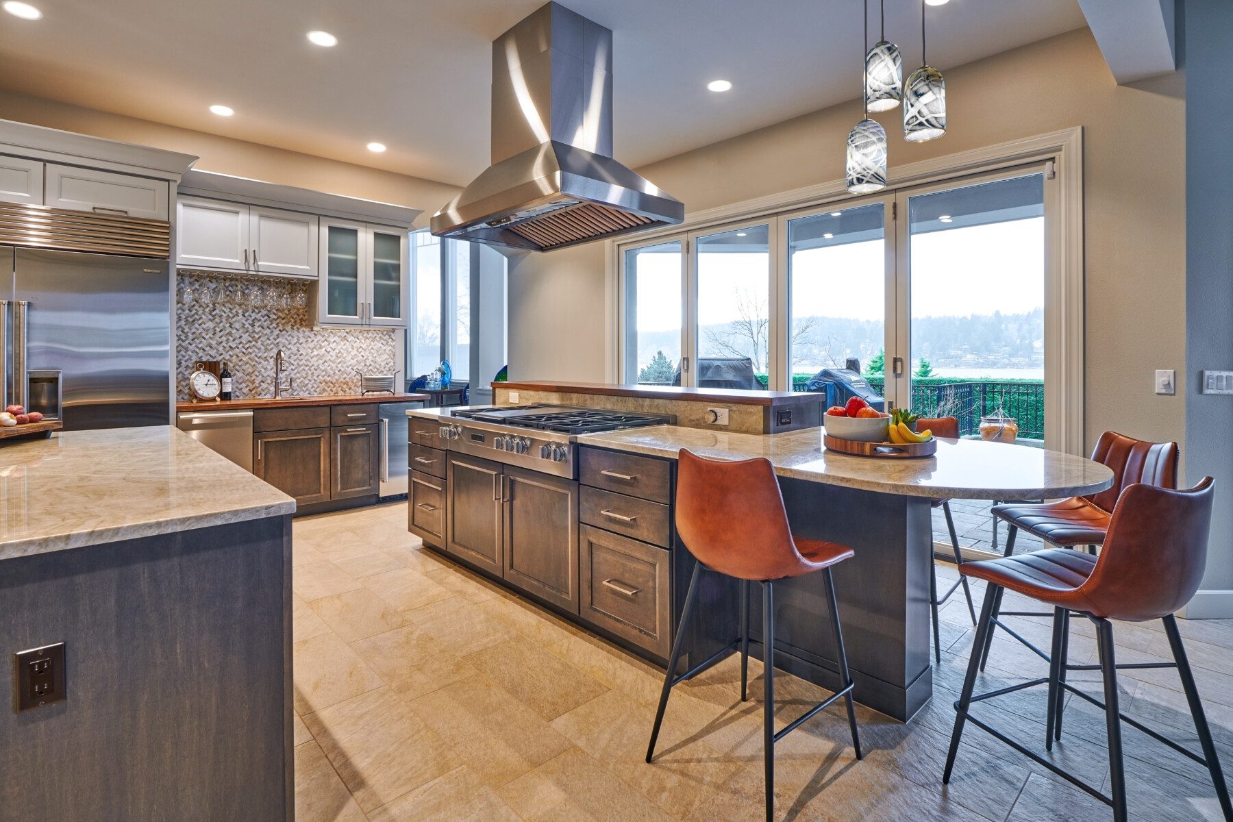 Modern kitchen with marble countertops, stainless steel appliances, a large range hood, bar seating with brown chairs, and large windows overlooking an outdoor patio.