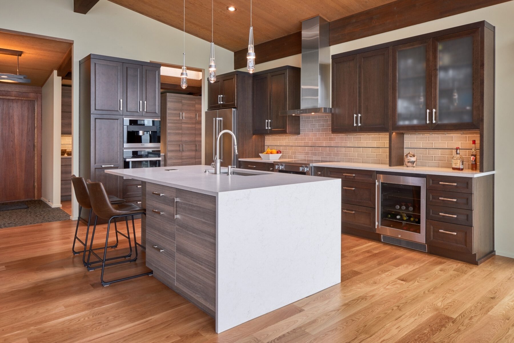 Modern kitchen with dark wood cabinets, a white island with a sink, built-in appliances, pendant lights, and two barstools on a light hardwood floor.