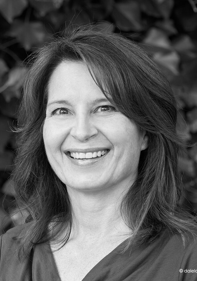 A woman with shoulder-length hair smiles at the camera in front of leafy foliage; the photo is in black and white.