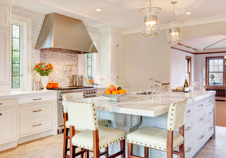 Modern kitchen with white cabinets, a marble island with seating, stainless steel range hood, pendant lights, and a bowl of oranges on the counter.