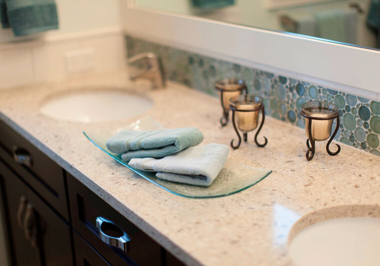 A bathroom countertop with two sinks, folded towels on a glass tray, three candle holders, and a circular tile backsplash.