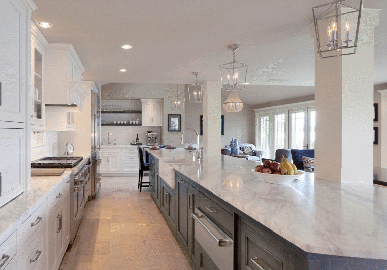 Spacious modern kitchen with white cabinets, marble countertops, pendant lighting, and a view into a living area with large windows. Fruit bowl on the central island.