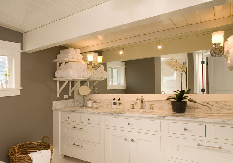 Modern bathroom with white cabinets, marble countertop, large mirror, rolled towels on shelves, a potted orchid, and a wicker basket on the floor.