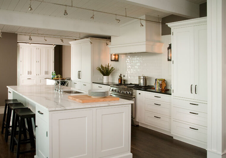 Modern kitchen with white cabinets, a large marble island with three black stools, stainless steel appliances, and wooden flooring.