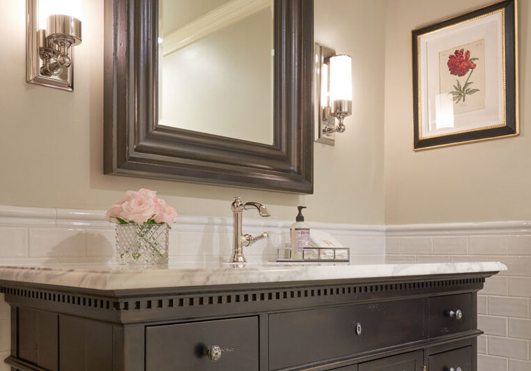 A bathroom vanity with a marble countertop, a framed mirror, wall sconces, a flower vase, soap dispenser, and a framed flower picture on the wall.