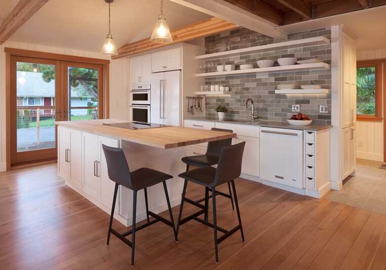 Modern kitchen with wood floors, white cabinets, a center island with two black chairs, open shelves, and gray tile backsplash. Large windows and pendant lights provide natural and artificial light.