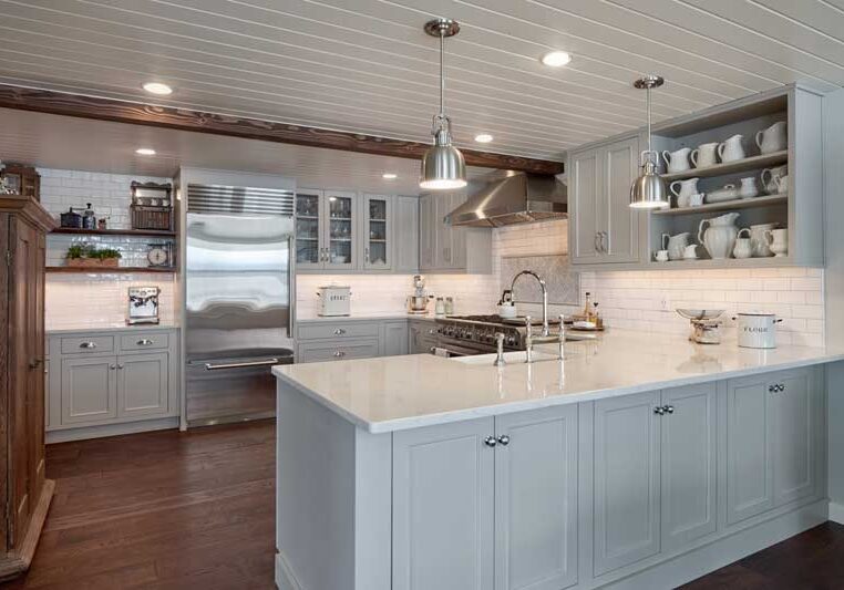 Modern kitchen with gray cabinets, a large white island countertop, stainless steel appliances, open shelving with dishes, and pendant lights over a dark wood floor.
