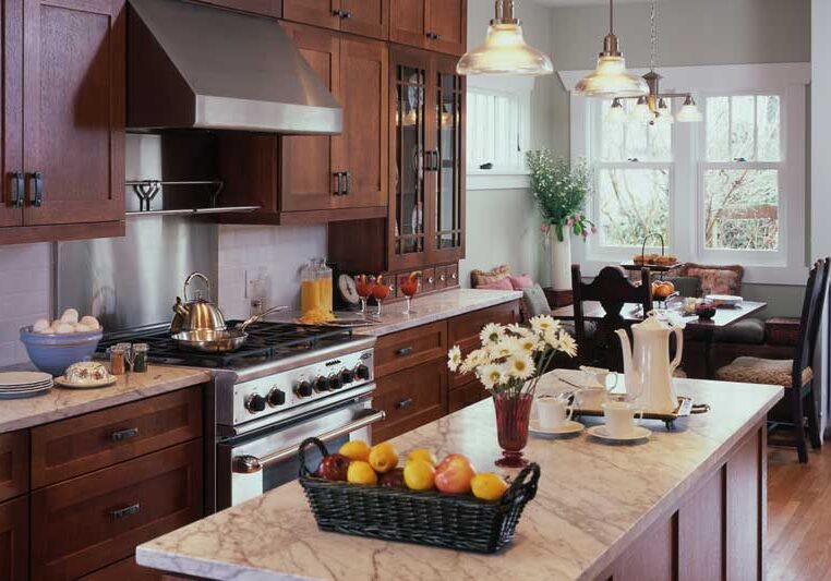 A modern kitchen with dark wood cabinets, marble countertops, stainless steel appliances, and a central island with a basket of fruit and a vase of white flowers.