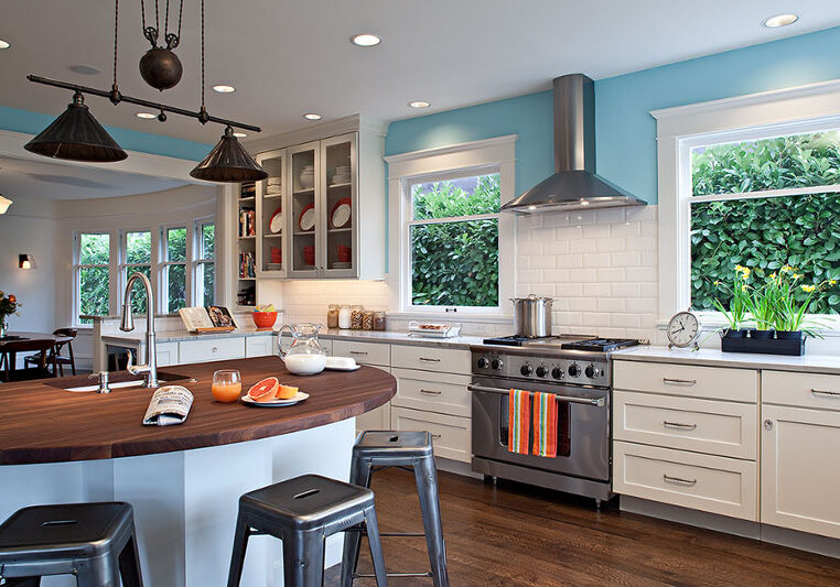 Modern kitchen with white cabinets, stainless steel appliances, a wooden island with stools, and large windows overlooking greenery. Cookware and dishes are visible on counters and shelves.