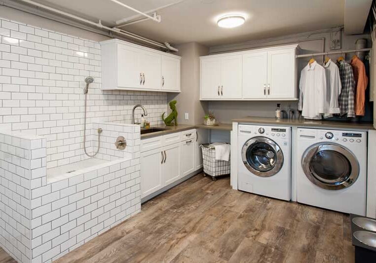 Modern laundry room with washing machine, dryer, white cabinets, tiled pet washing station, and clothes hanging on a rail. Wood-look flooring and countertop with laundry supplies.