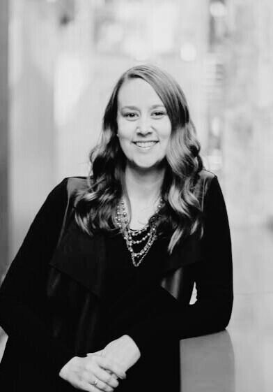 A woman with wavy hair, wearing a dark top and layered necklace, smiles while leaning on a surface in an indoor setting. The photo is in black and white.