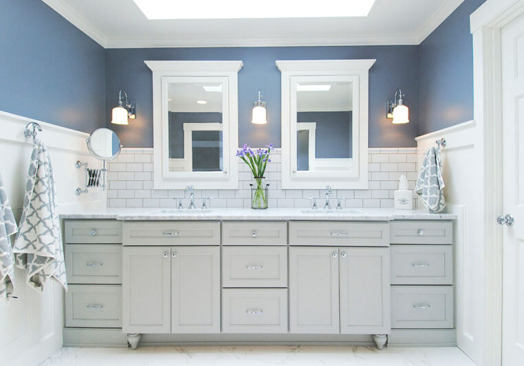 Modern bathroom with double sinks, gray cabinets, white subway tile backsplash, two mirrors, wall sconces, and a vase of purple flowers on the countertop.