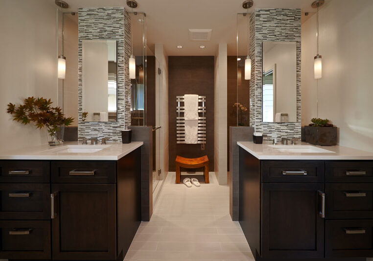Modern bathroom with double sinks, dark wood cabinets, mosaic tile accents, two pendant lights, a walk-in shower, towel warmer, and decorative plants on each countertop.