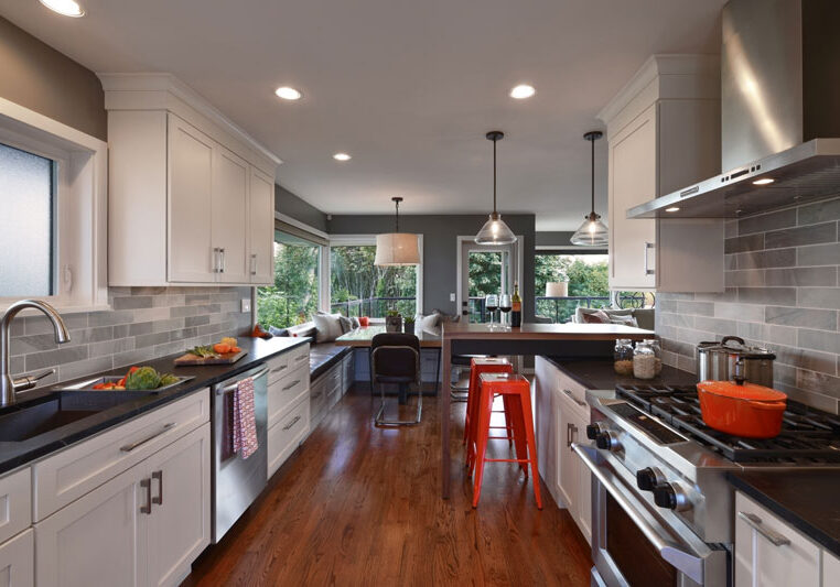 Modern kitchen with white cabinets, gray backsplash, stainless steel appliances, a breakfast nook with large windows, and wooden flooring. Orange barstool and cookware add color accents.