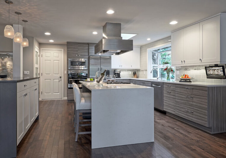 Modern kitchen with white and gray cabinets, stainless steel appliances, a large island with stools, wood flooring, and pendant lighting. Fruit and decor are on the counters.